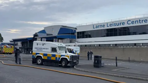 PA Media Two white and yellow police vans sit in front of a large grey and black building with the the words 'Larne Leisure Centre' in blue font on it. To the left of the frame a red and yellow fire truck is parked. Three police officer in black riot gear and black helmet hold clear riot shields.