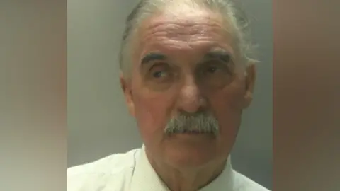 A man with grey hair and moustache. He is wearing a white shirt. 