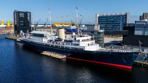 The Royal Yacht Britannia moored at Leith, with its dark blue hull, white upper decks, signal‑flagged masts, and a modern distillery building in the background under a clear blue sky