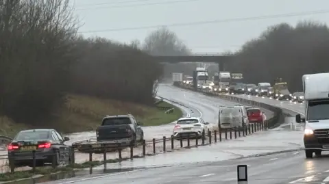 Flooding on the A30 near Ottery St Mary. There are cars and long tailbacks on the left side of the road, and cars in flood water on the opposite side.