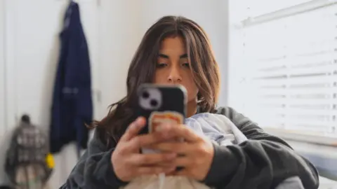 Getty Images. Una joven usa su teléfono inteligente mientras se prepara para lavar la ropa.