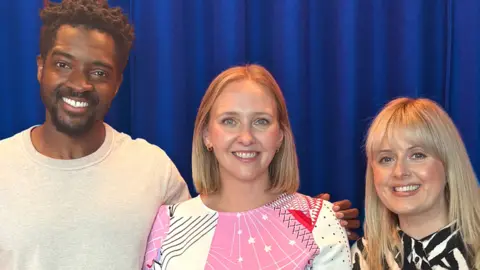 Three smiling adults posing together in front of a blue curtain