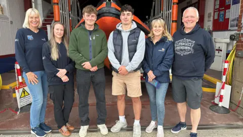 The two brothers stand with four RNLI volunteers, three women and a man who are wearing navy RNLI jackets. Behind them is the lifeboat.