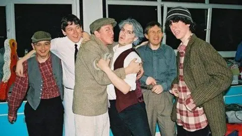 Peter Wicks Peter and his friends pose for a photo dressed up for a high school drama production. They wear tartan shirts, waistcoast, flatcaps and make up. 