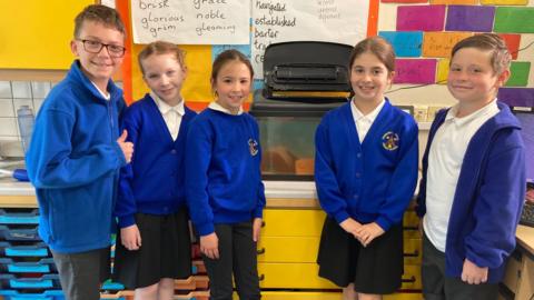 Five pupils at Churchill School in Langford, three girls and two boys pose in front of the eel tank wearing blue jumpers.