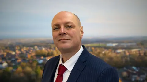 Great Dawley Town Council A bald man wearing a blue pin-stripped blazer, white shirt and red tie, standing in front of an aerial image of a blurred town