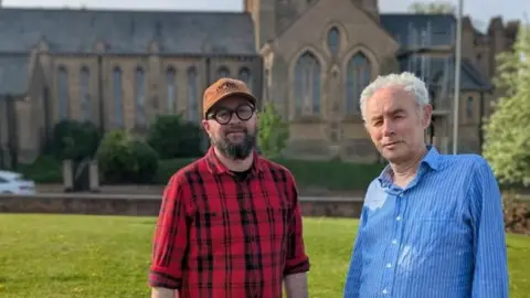 Narbi Price Artist Narbi Price (left) and Mark Hudson are standing on grass in front of the church. Mr Price is wearing a cap, glasses and a red checked shirt. He has a dark beard and moustache. Mr Hudson is in a blue shirt, with small checks, and has short-ish white hair.