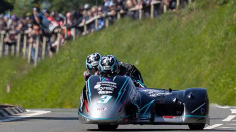 A dark coloured sidecar outfit on a race track with a crowd looking on