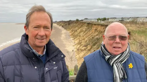 GUY CAMPBELL/BBC Clive Crossley, wearing a blue jacket, and John Pitts, wearing a scarf and glasses, stand on a cliff edge with eroded beach in background  