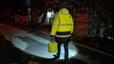 PA Media A person dressed in a yellow high vis jacket walking down a dark lane. They are holding a torch and shining it on the road. They are carrying a bucket.