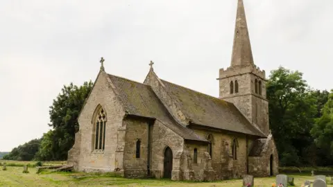 A church with a spire. It has a churchyard, and there are trees on the perimeter.