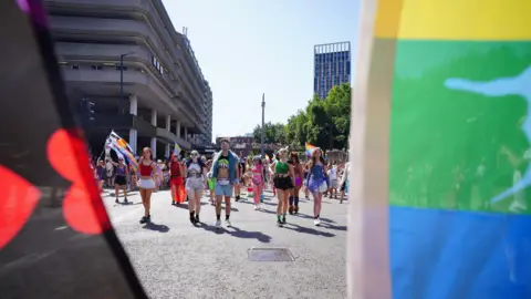 PA Media Shot looking between two flags down a city road with people walking in the march - very much dressed for the heat in shorts