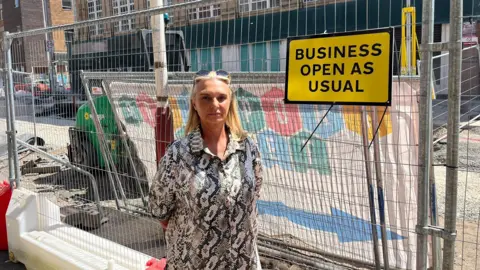 Julie Goodwin has long blonde hair and has sunglasses on top of her head. She is wearing a grey and white patterned shirt and is standing in from of metal roadworks fencing. There is a yellow sign with black writing which reads Business open as usual.