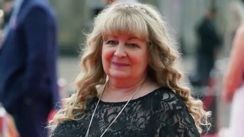 PA Media comedian Janey Godley with long blonde hair and a headband smiling at the camera. She wears a black lace top and a long silver necklace.