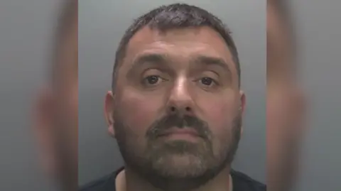 Lee Kent mugshot against a grey background. He has a short beard and short black hair