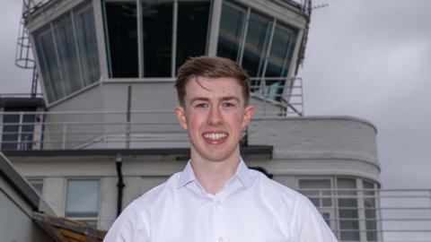 Ben Harrety in front of Teesside Airport control tower.  He's wearing a white shirt and is smiling.