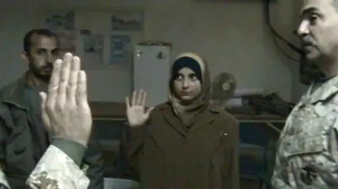 US Marine Corps A teenage Safa holds up her right hand and looks at a man in front of her. She is wearing a headscarf and khaki overcoat.  There are three men in the image who can only be partly seen - two are in camouflage clothing. The one she is looking at is also holding up his right hand. 