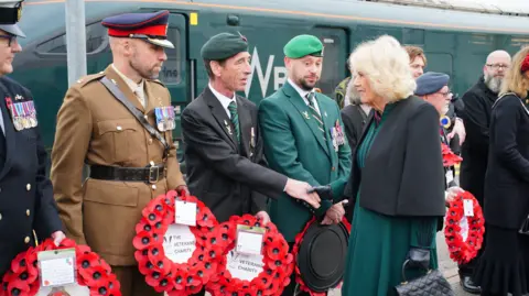 The Queen shakes hand with a smartly dressed veteran, who stands in a line of other veterans holding wreaths and many in uniform. All are in front of a dark green GWR train.
