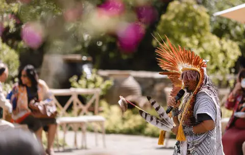 Courtney Louise/The King’s Foundation A man wearing traditional dress holds two black and white feathers and a microphone at Highgrove
