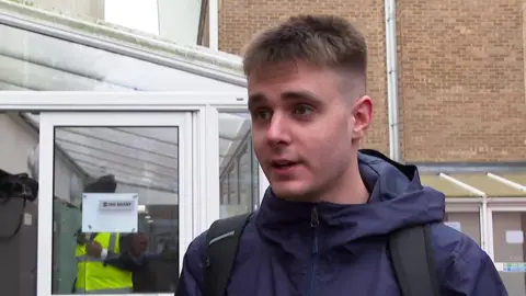 A head and shoulders image of James Thompson. He is wearing a navy coat over which he has two black rucksack straps. He is looking directly past the camera and standing in front of a conservatory-like entrance to a university building.