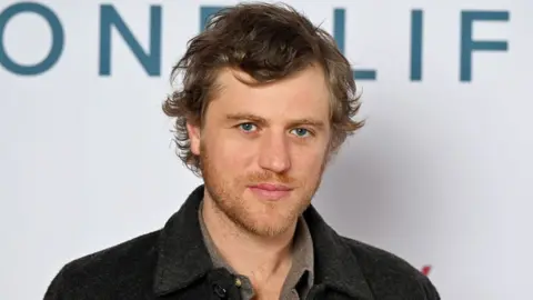 Getty Images Johnny Flynn standing looking at the camera against a white backdrop at a film premiere