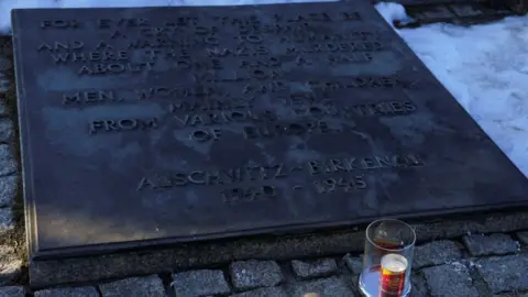 Reuters A candle in a clear holder is burning in front of a plaque at Auschwitz-Birkenau, which is surrounded by snow.