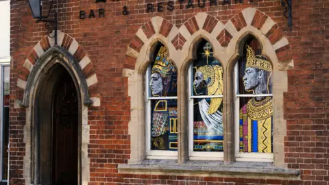Ash Field/Our Tiny Windows A brick building with arched windows and an arched door has the words 'bar & restaurant' written at the top. The three windows are painted with figures covered in gold, blue and red colours. They appear to the the three wise men/three kings.