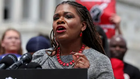 Cori Bush in grey suit with red necklace and earrings at lectern with microphones