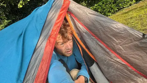 Ollie White Ollie, looking tired as though he has just woken up, pokes his head out of a blue, grey and red tent.