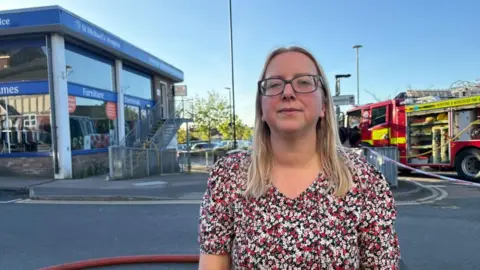 A woman in a floral patterned dress with blond hair and glasses is standing on the corner of a road. Behind her is the corner of a commercial building with a metal staircase heading up the side of it. There is a fire engine, in use on the other side of the street.