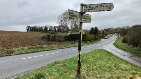 A T-junction in the countryside. It is a dark and overcast day and the road is wet, surrounded on either side by grassy verges, ploughed fields and bushes lining the road. There is a road sign in the middle of the picture on a grassy triangle, pointing in the directions of Fairford, Cirencester and Burford.