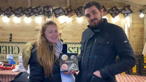 Two market traders in their wooden chalet smiling for the camera. A man, located on the right of the image, has dark hair and a beard. He is wearing a black coat, and his hands are in his pocket. The woman, on the left of the image, is wearing a grey hoodie and has long brown hair. 