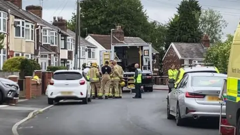 Emergency services in a residential street gathered in front of emergency vehicles - in between residents' parked cars - as they deal with the discovery of explosives in a home in St Helens.
