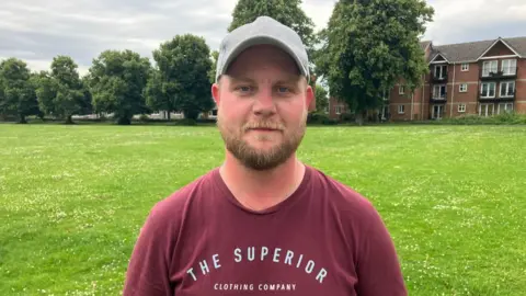 Rhys looks at the camera. He is wearing a burgundy t-shirt and has a grey baseball hat on. He has a short sandy-coloured beard. Grass can be seen behind him with a block of flats in the background.