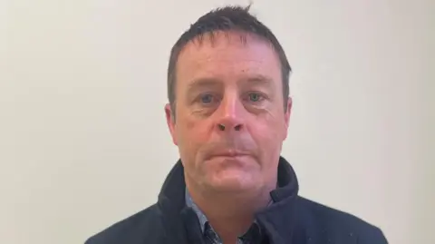 A man with short brown hair looks into the camera. He is wearing a dark overcoat with the collar turned up. He is standing in front of a light coloured wall.