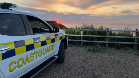 Cromer Coastguard Rescue Team A coastguard vehicle parked, looking out towards the sea as the sun sets, casting a vibrant orange and purple hue across the North Sea.