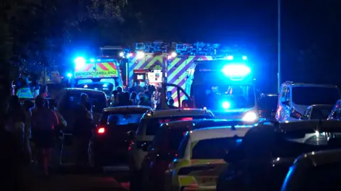 Scene of the road after the collision. Several ambulances and parked cars, and police cars litter the road. It is dark. Several people mill around the cars.