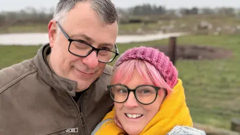 Katie Brown Two people are standing close together outdoors, with one resting their head against the other. The man on the left is wearing a brown jacket with a front zip and a visible zippered pocket. The woman on the right is wrapped in a thick yellow scarf and is also wearing a pink knitted hat and a grey-and-blue textured coat. The background shows an open, grassy landscape on an overcast day.
