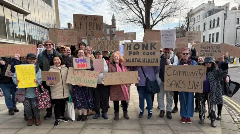 A group of people stand outside a building on a street. They are holding home-made placards in support of a mental health service which risks being cut. 