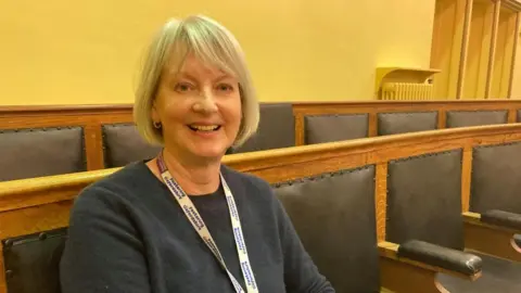 A woman is sat on a wooden bench with yellow walls behind her. She is smiling and wearing a lanyard.