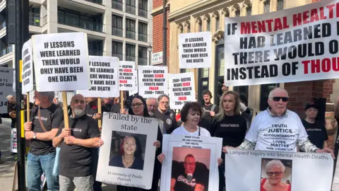 Richard Daniel/BBC Bereaved families standing outside the inquiry building, holding posters of their relatives. They are also holding placards saying things like 'if lessons were learned, there would have only been one death' and 'failed by the state'. There are at least 50 families standing in protest outside the inquiry. About 20 can be seen in this image.