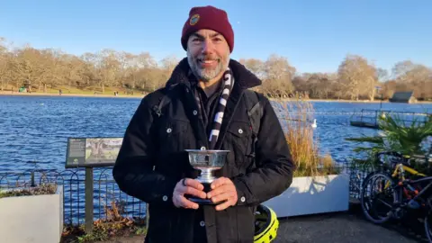 A smiling man holds a trophy. behind him is the Serpentine in London's Hyde Park. 