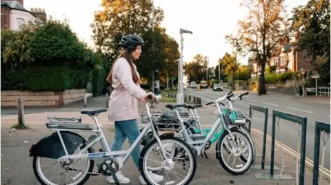 A woman wearing a cycle helmet and light pink jacket parks up her ebike on a street station along a residential road lined with trees and elegant terraces on the opposite side.