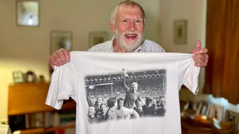 A man with silver hair grins at the camera as he holds up a t-shirt with the famous black and white image of England captain Bobby Moore holding the World Cup trophy aloft, hoisted up in the air by his teammates, after the team won the 1966 tournament.