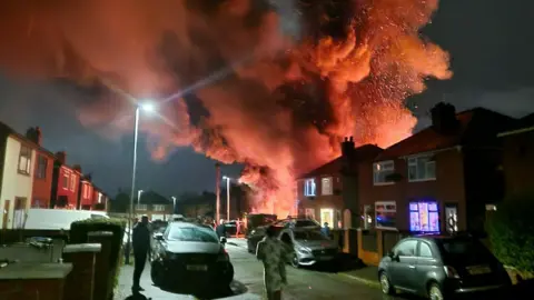 Billowing smoke and flames as the fire burned at the house. People are hurrying around near the house and cars are park on both sides of the road.