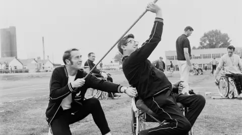 Black and white photo. A man - Jim Savage - sits in a wheelchair leans backwards with one arm raised up holding a javelin. Another man kneels on the ground behind him and holds the end of the javelin while holding the wheelchair steady. Other wheelchair users are dotted around the field. In the background on the left are the huts, which are pre-fab and lined up together. 19th International Stoke Mandeville Games at the Stoke Mandeville Stadium in Aylesbury, Buckinghamshire, UK, 17th July 1970, His team-mate Ross MacFarlane lends support to one of the wheels on his wheelchair. 