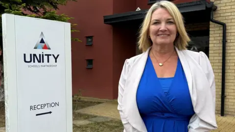 USP Sarah Garner wears a blue dress and white jacket and smiles. She is standing in front of a sign saying Unity Schools Partnership.