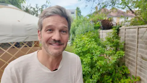 A man in a light coloured t-shirt with grey hair and a beard stands in a back garden with shrubs and a yurt in the background