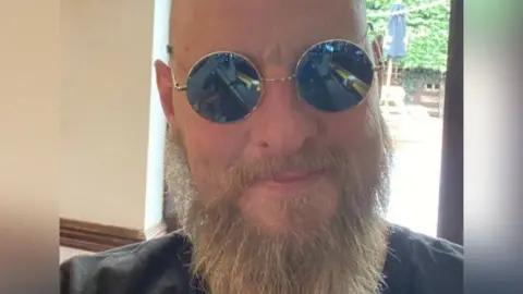 Handout A close-up of Paul Hart, who is bald and has a long beard and is wearing sunglasses. He is in front of a window and a parasol and garden furniture can be seen outside.