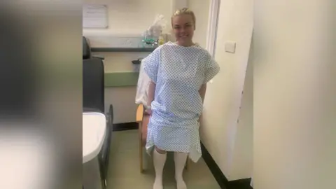 Ellie Philpot A blonde girl, Ellie, is in a hospital gown with a small square pattern is standing in a hospital room smiling. She had long white socks on and looks to be preparing for an operation.